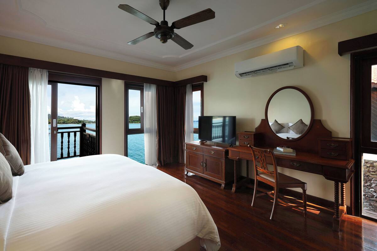 Ein Hotelzimmer mit einem großen Bett, einem Schreibtisch mit Fernseher, einem Deckenventilator und Blick auf das Meer.