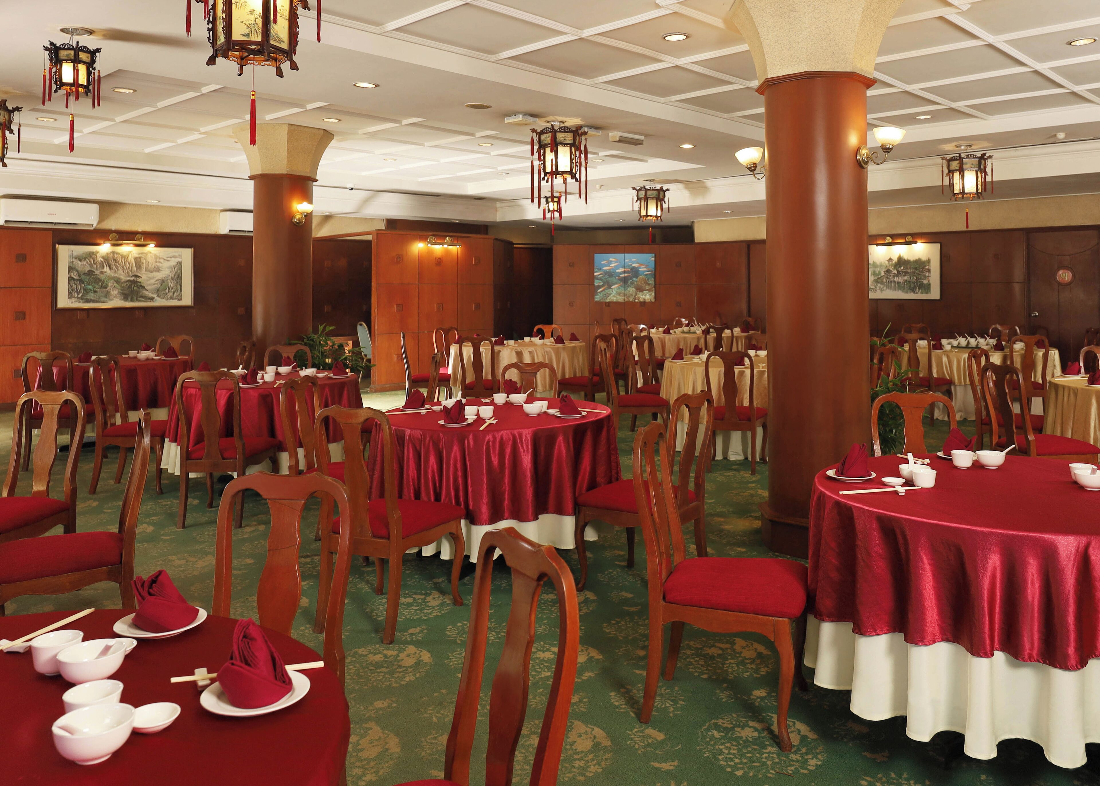 A banquet hall with round tables covered in red tablecloths, wooden chairs, and hanging lanterns.