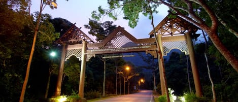 A wooden archway with intricate lattice work, leading to a road flanked by trees and illuminated by street lamps.