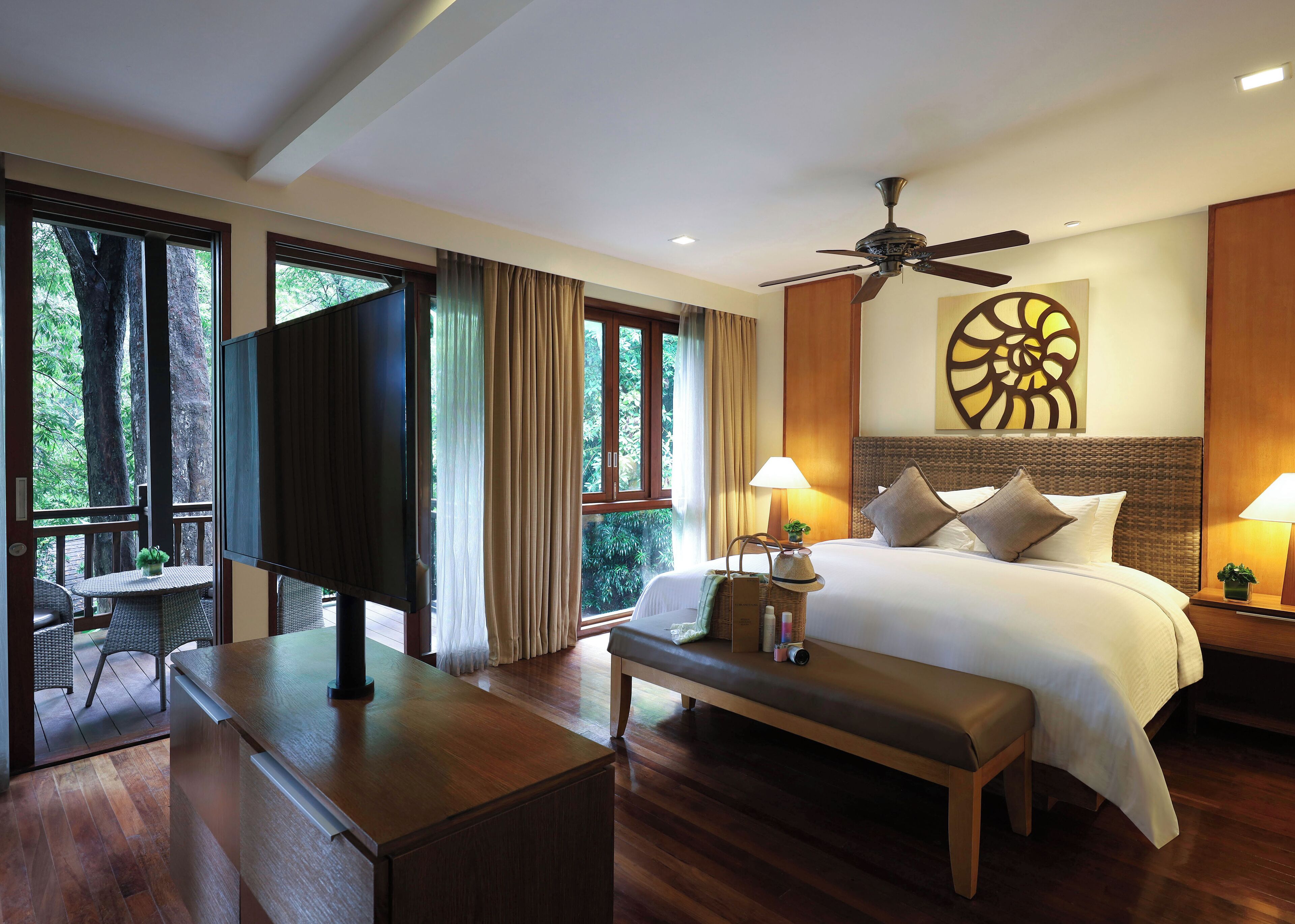 A hotel room with a bed, bedside table, lamp, and a view of trees through large windows.