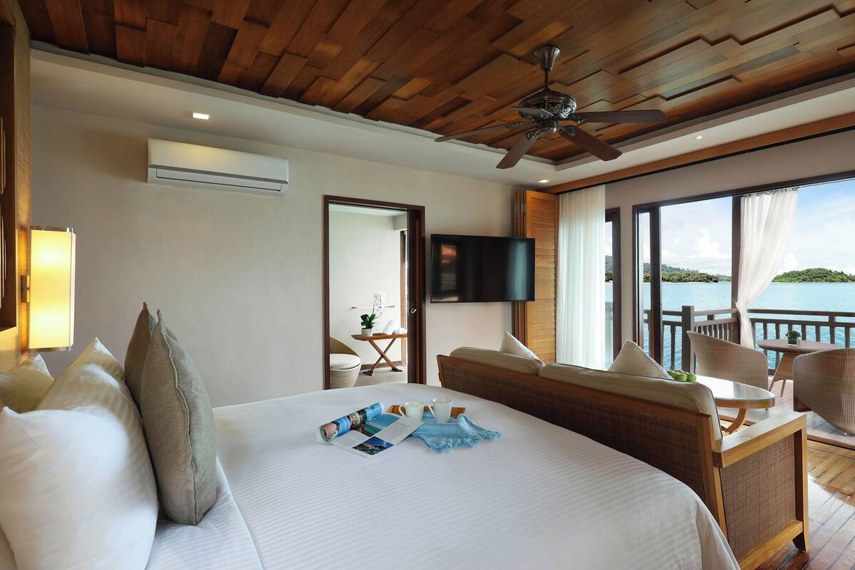 Ein Hotelzimmer mit einem großen Bett, einem Deckenventilator, einem Fernseher und einem Balkon mit Blick auf das Wasser.