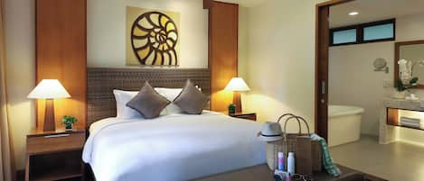 A hotel room with a large bed, two bedside tables with lamps, a ceiling fan, a wall art piece, a bathroom with a bathtub, and a bench with toiletries.