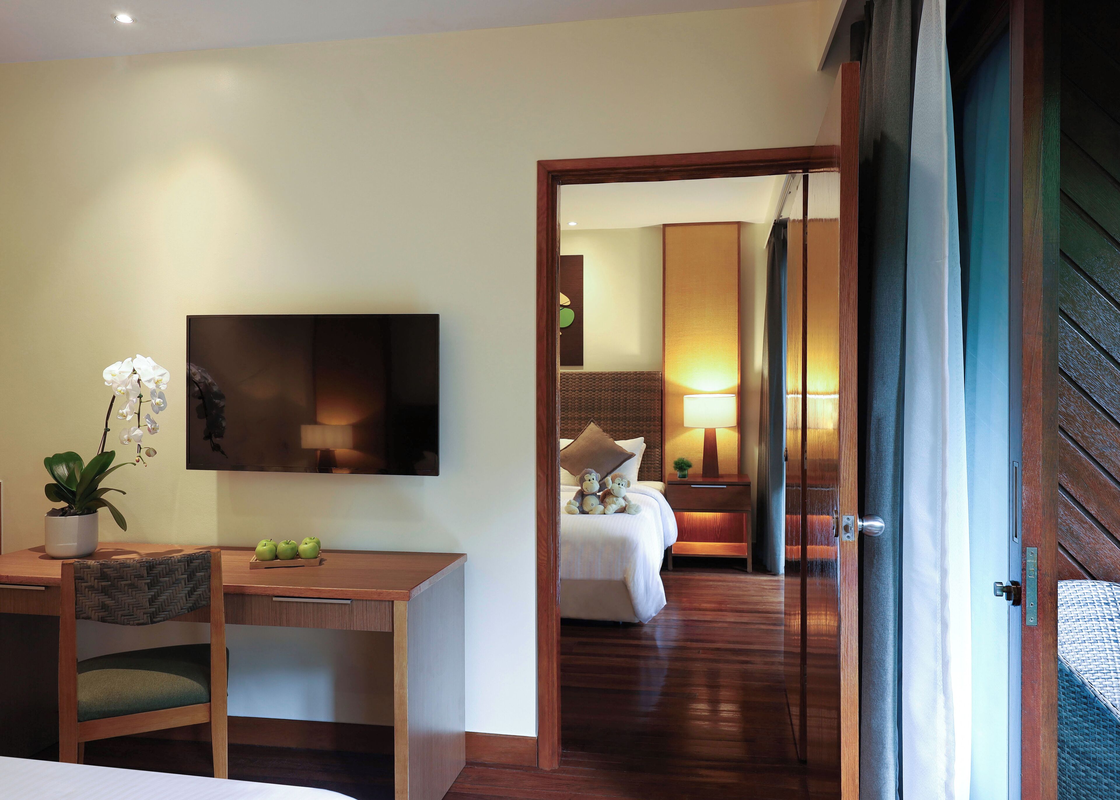 A hotel room with a bed, a desk, a chair, a television, a lamp, and a door leading to another room.