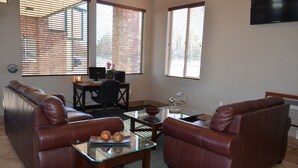 A living room with leather sofas, a glass coffee table, a desk with a computer, and large windows with blinds. - National 9 Price (Price)