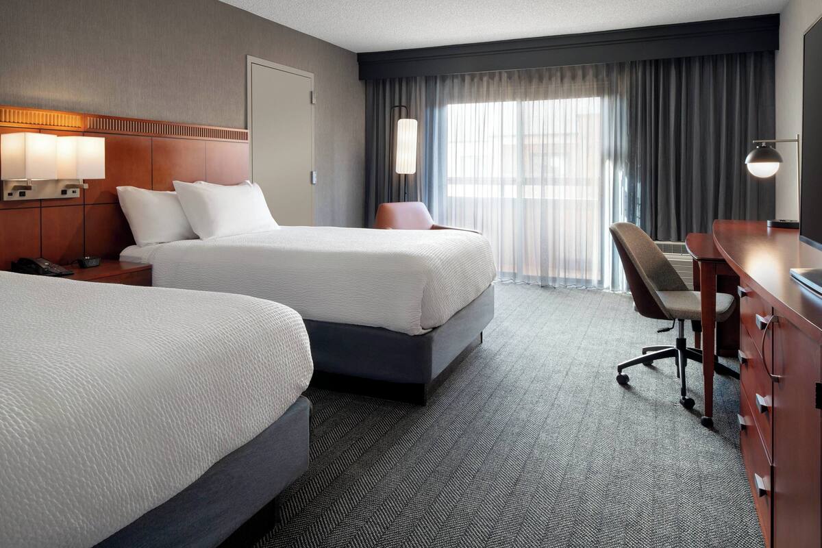 A hotel room with two beds, a desk, a chair, and a large window with curtains.