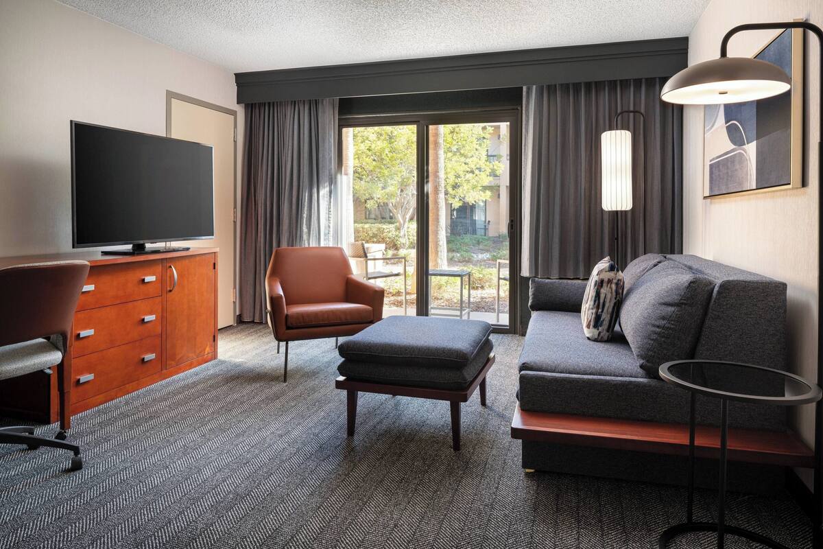 A hotel room with a flat-screen TV, a wooden cabinet, a leather armchair, a grey sofa, a small round table, and a large window with curtains.