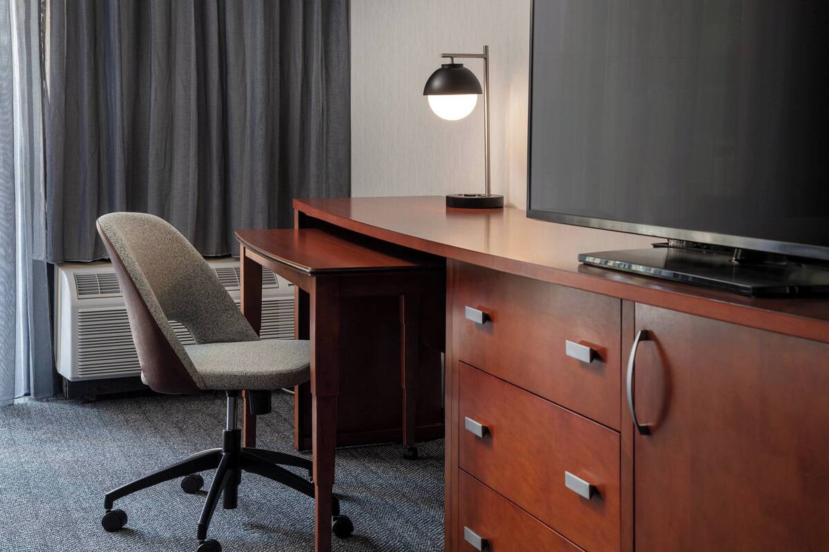 A hotel room with a wooden desk, a modern chair, a flat-screen TV, a desk lamp, and a large window with curtains.