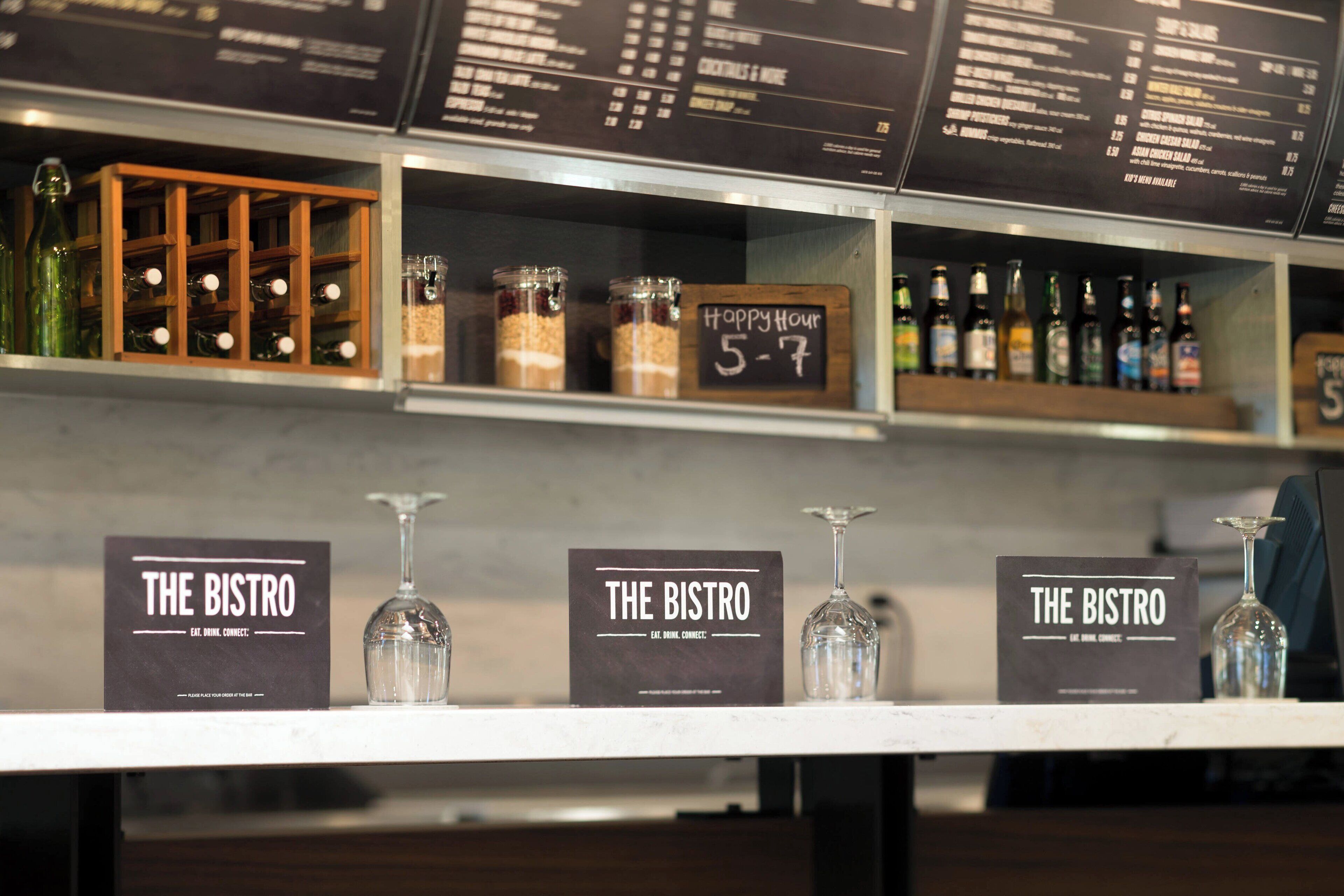 Uma área de bar com prateleiras exibindo garrafas e copos. Há placas com a inscrição 'The Bistro' e um aviso de 'Happy Hour'.