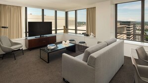 A modern hotel room with a large window offering a city view, a flat-screen TV, a sofa, a coffee table, and a desk with a chair. - Hilton Portland Downtown (Portland)