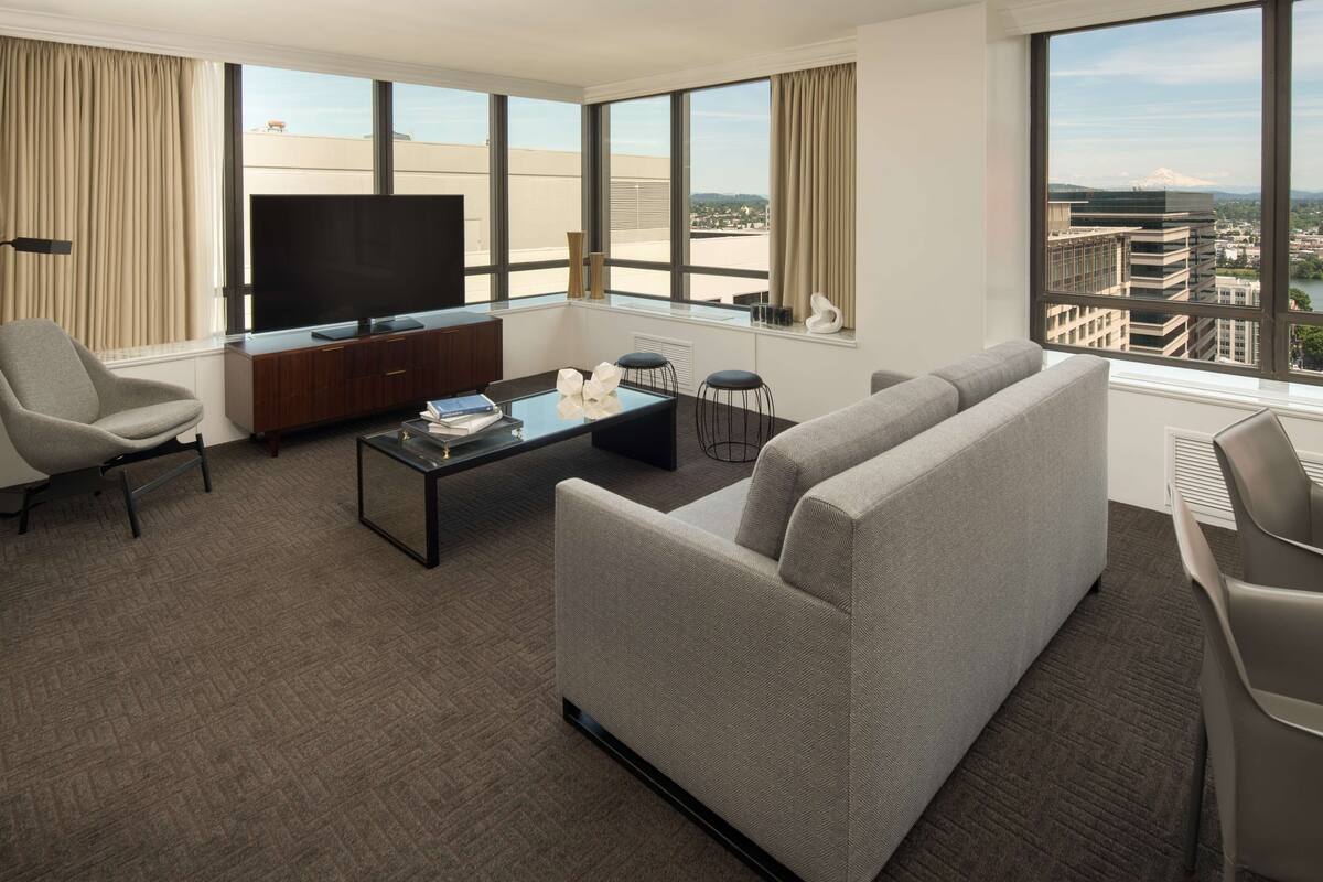 A modern hotel room with a large window offering a city view, a flat-screen TV, a sofa, a coffee table, and a desk with a chair.