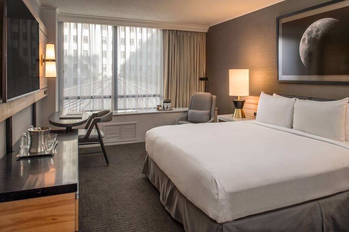 A hotel room with a large bed, a desk with a lamp, a chair, and a window with curtains.
