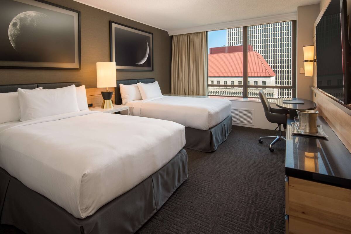 A hotel room with two beds, a desk, a chair, a TV, and a window with a city view.