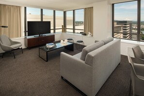 A modern hotel room with a large flat-screen TV, a sofa, a coffee table, and a view of the cityscape. - Hilton Portland Downtown (Portland)