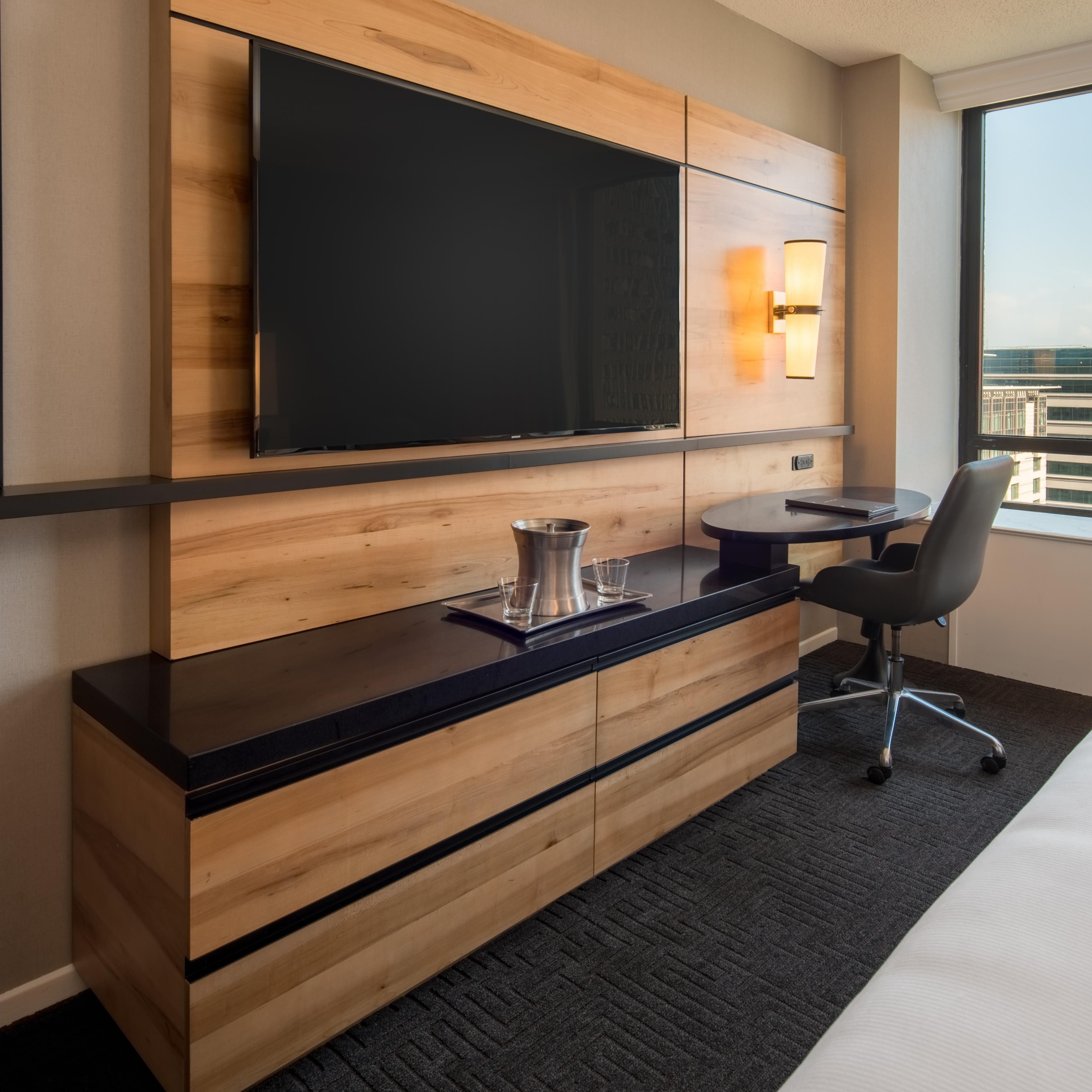 Habitación de hotel con televisor de pantalla plana montado en un panel de madera, una mesita con una silla y una ventana con vistas a edificios.