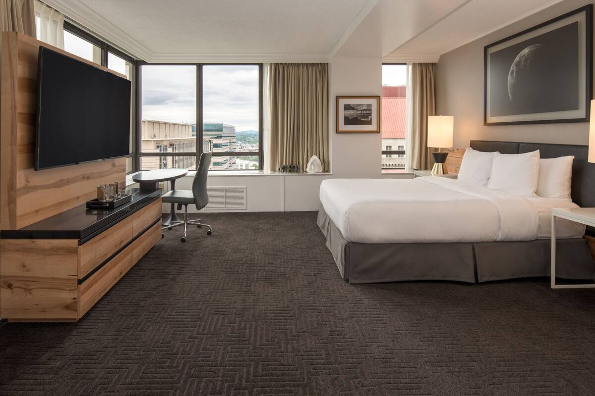 A hotel room with a large bed, a desk with a chair, a flat-screen TV, a window with a view of the city, and a framed picture on the wall.