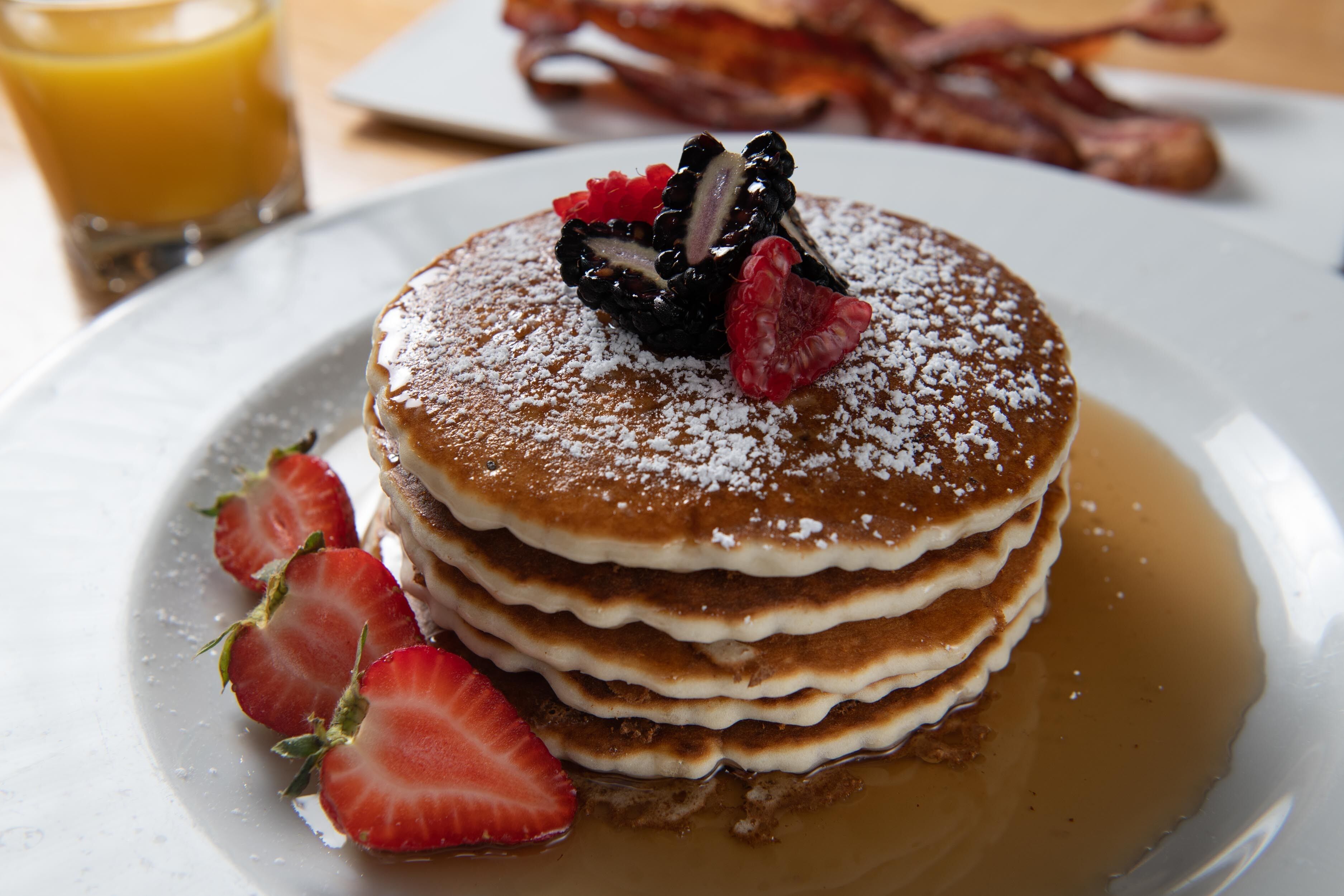 Una pila de tortitas con fresas y un recipiente con almíbar.