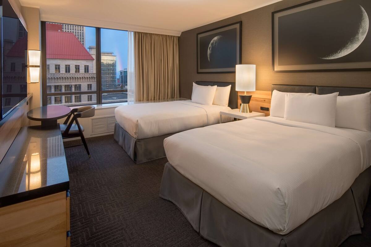 A hotel room with two beds, a desk, and a large window offering a city view.