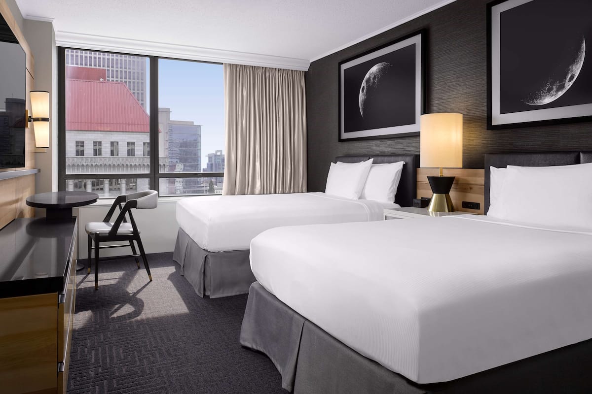 A hotel room with two beds, a desk, a chair, and a large window offering a city view.
