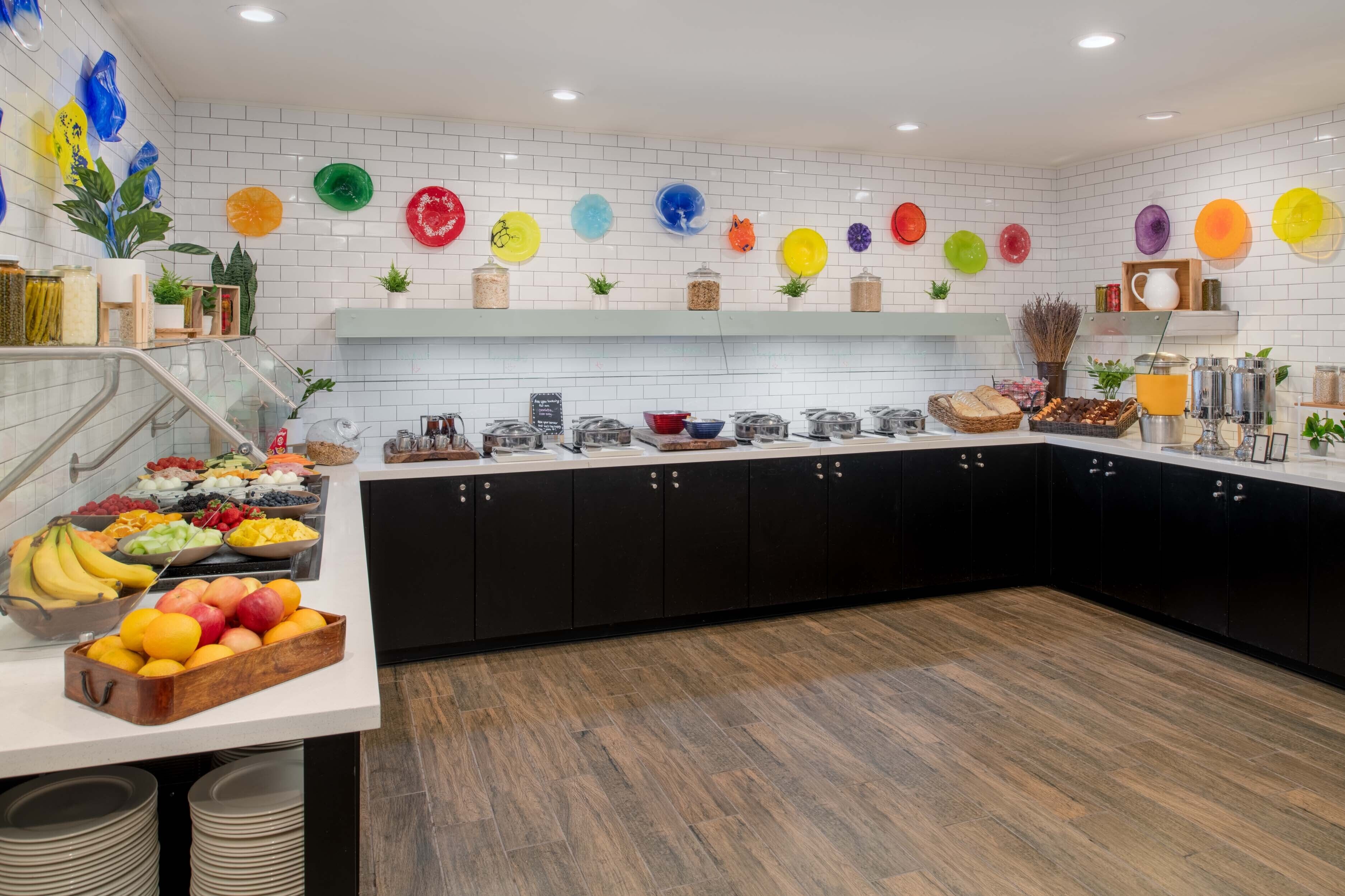 A modern kitchen with white tiled walls, black cabinets, and a variety of fruits and breakfast items on the counter.