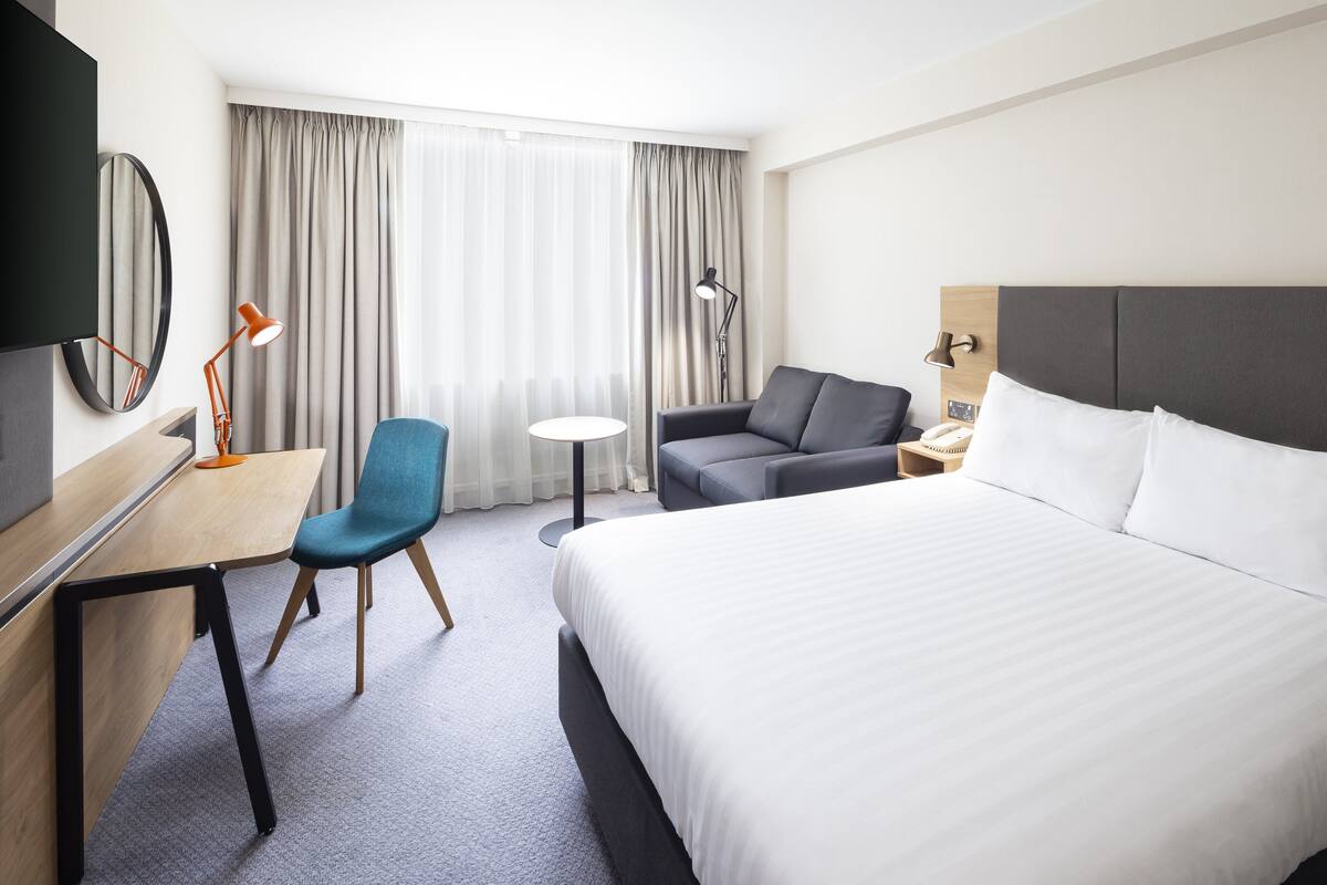 A hotel room with a bed, a desk with a chair, a sofa, a small round table, and a TV mounted on the wall.