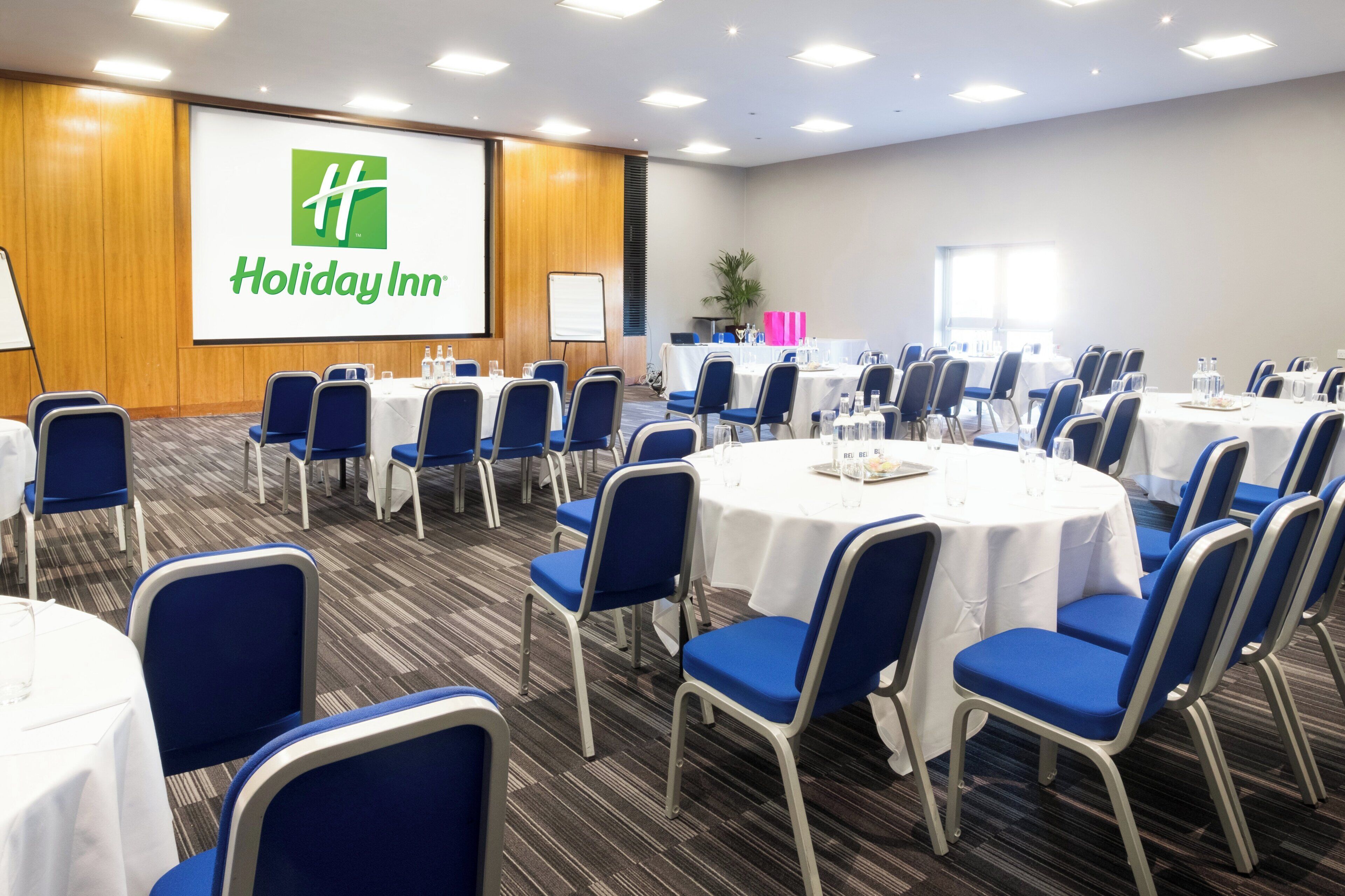 A conference room with round tables, blue chairs, and a projection screen displaying the Holiday Inn logo.