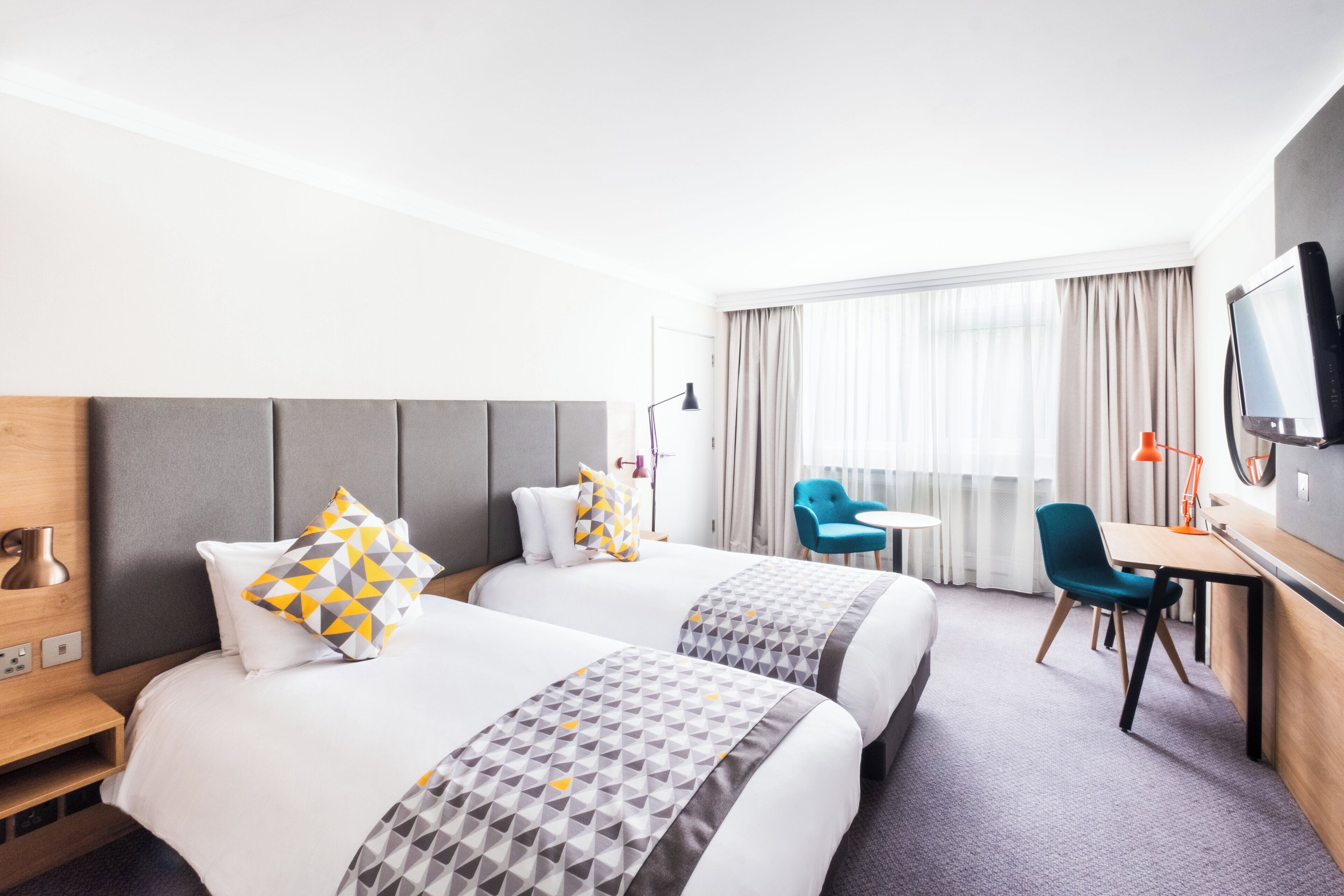 A hotel room with two beds, a desk, a chair, a television, and a large window with curtains.