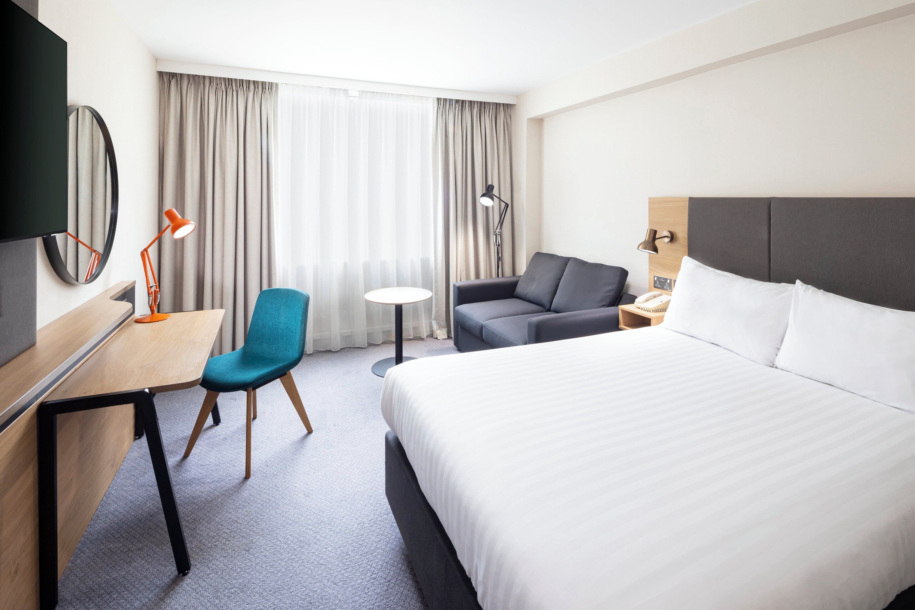A hotel room with a bed, a desk, a chair, a sofa, a TV, and a window with curtains.