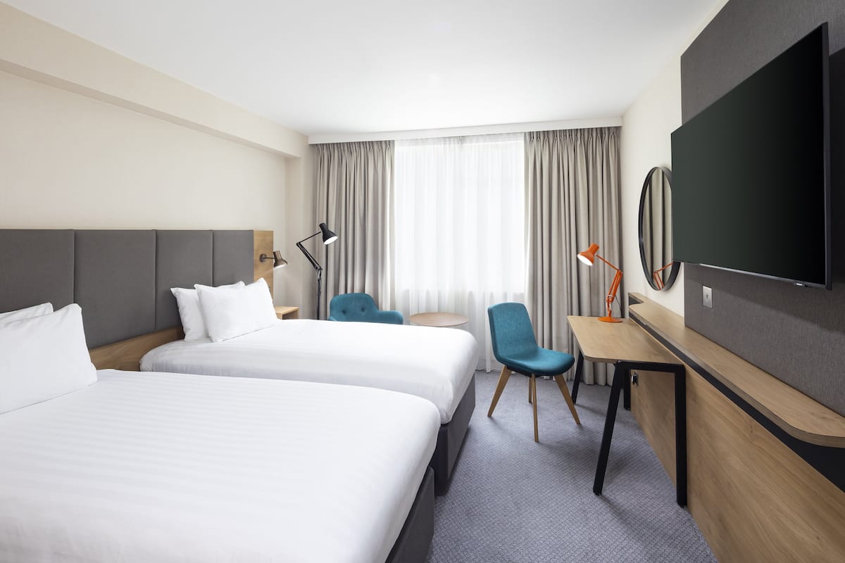 A modern hotel room with two beds, a desk, a chair, a TV, and a large window with curtains.