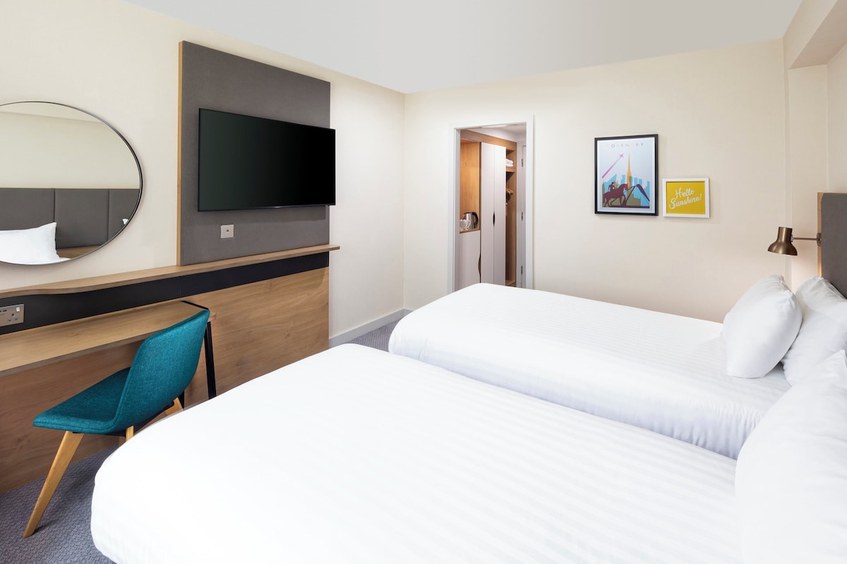 Habitación de hotel con una cama, un escritorio con silla, un televisor y una puerta que da a otra habitación.