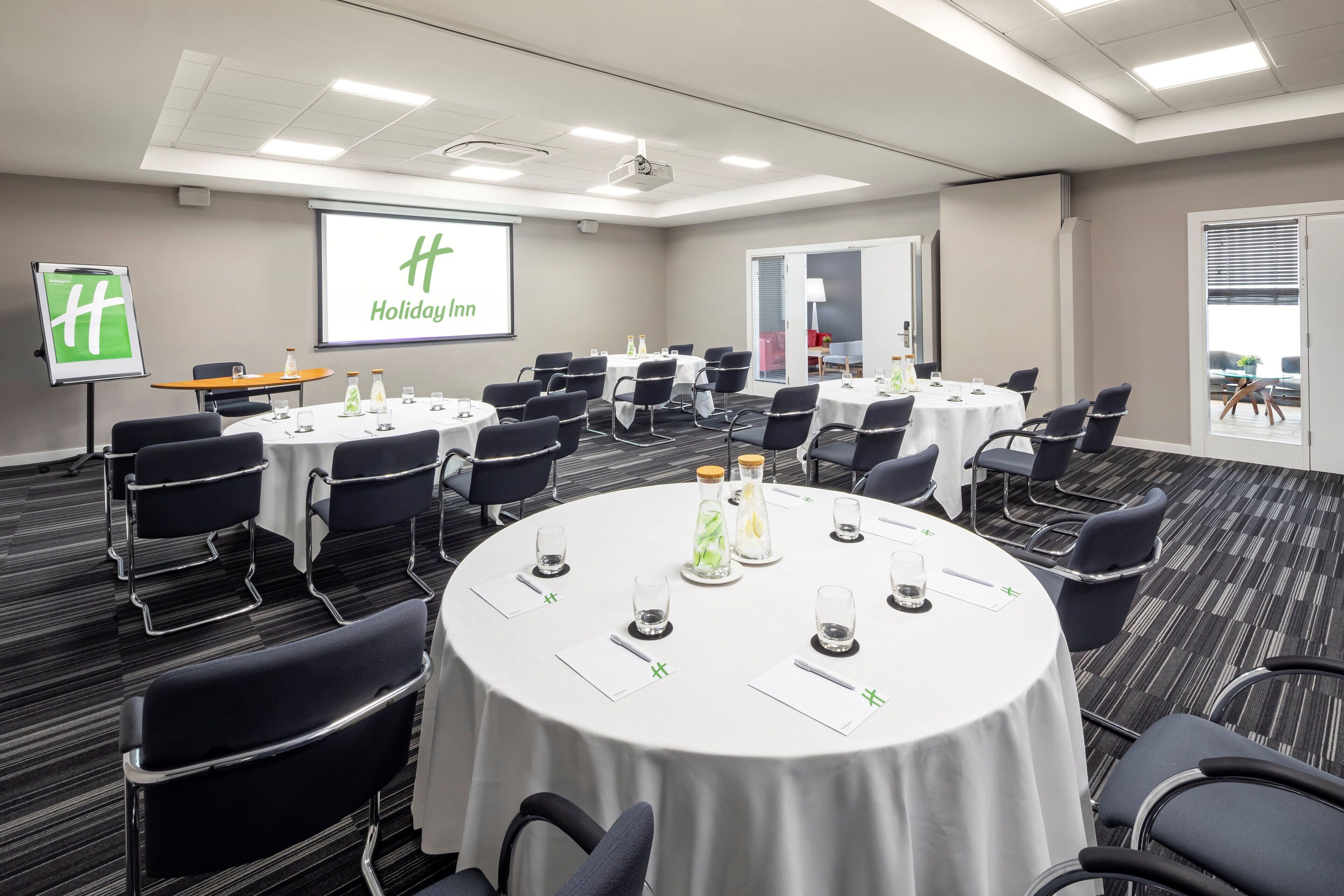 A conference room with multiple tables, chairs, and a projector screen displaying "Holiday Inn".