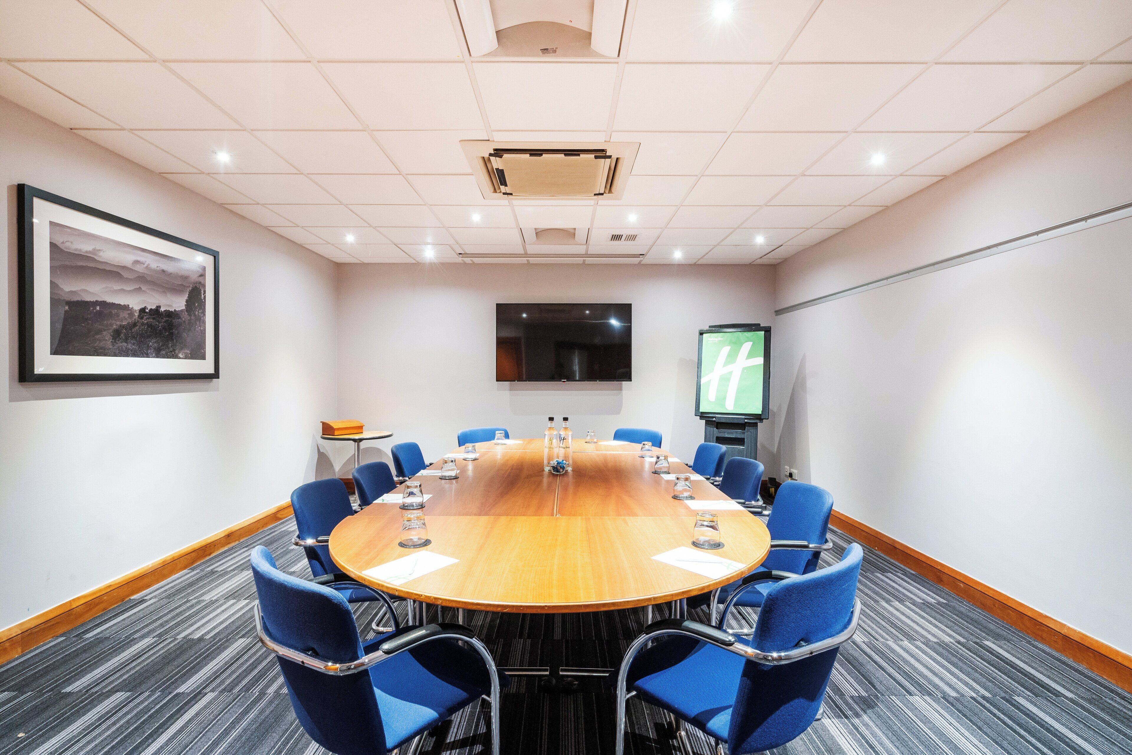 A conference room with a long table, blue chairs, a projector screen, and a framed picture on the wall.