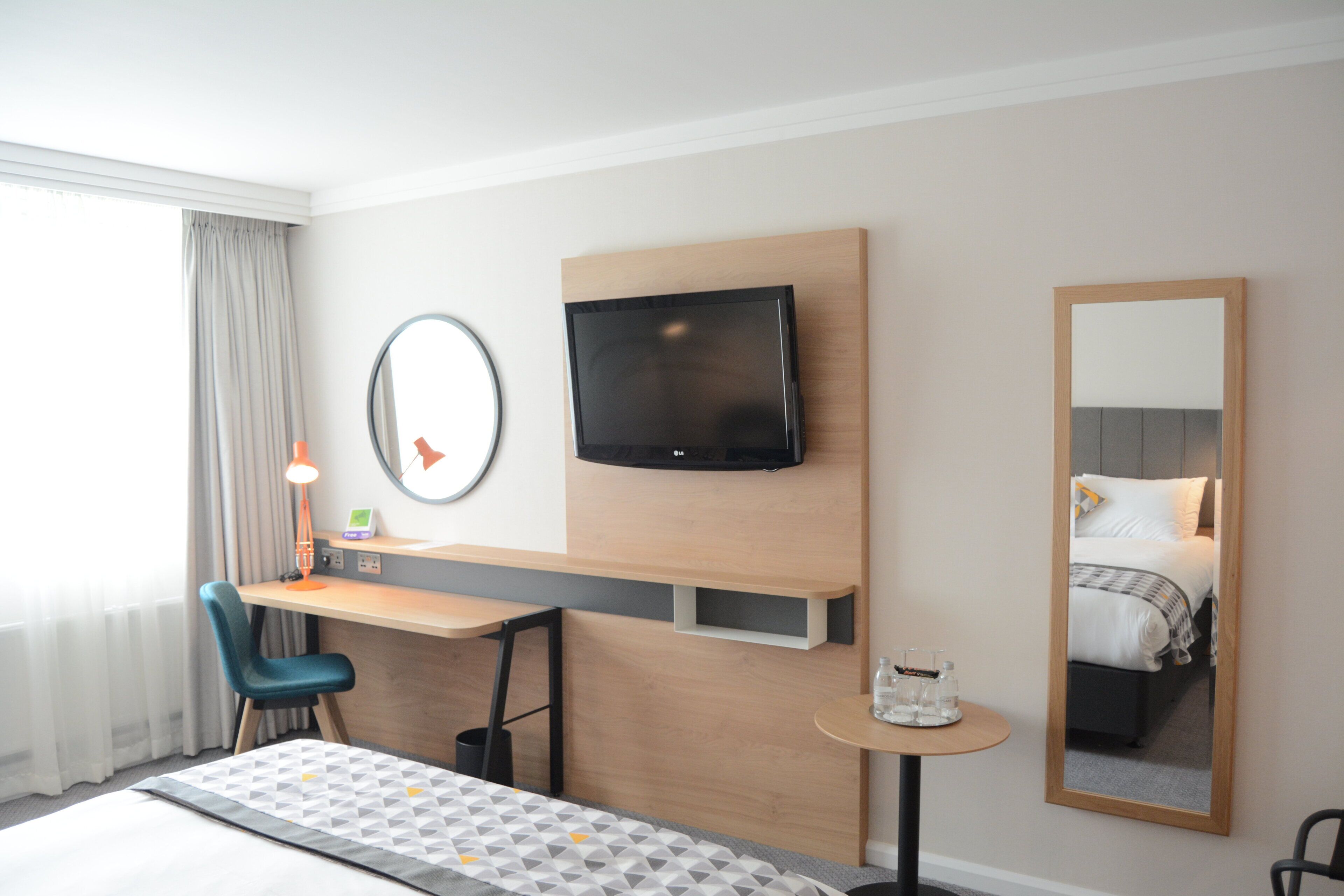 A hotel room with a bed, a desk with a chair, a television, a small table with a mirror, and a window with curtains.