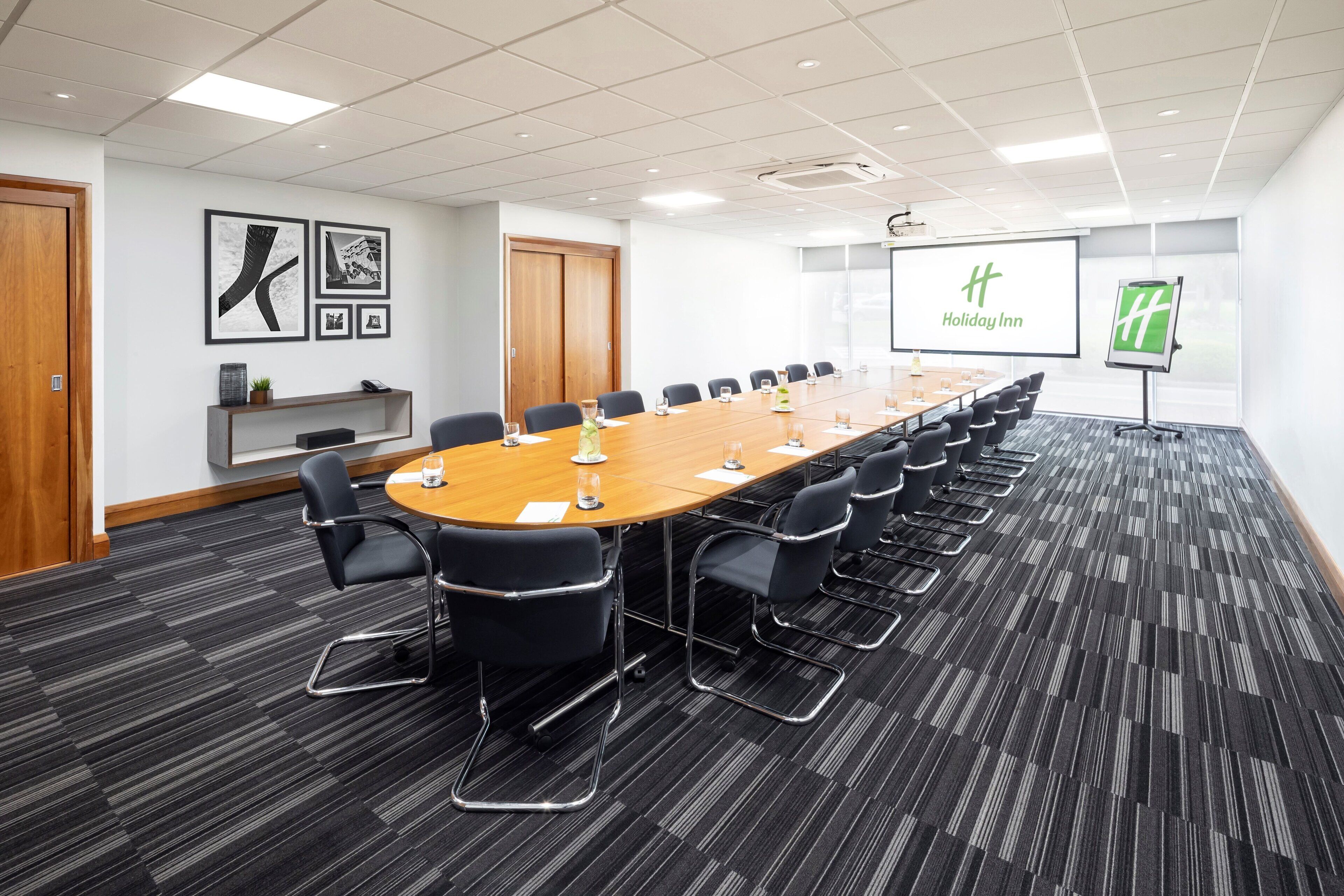 A conference room with a long table, black chairs, a projector screen, and a Holiday Inn logo.