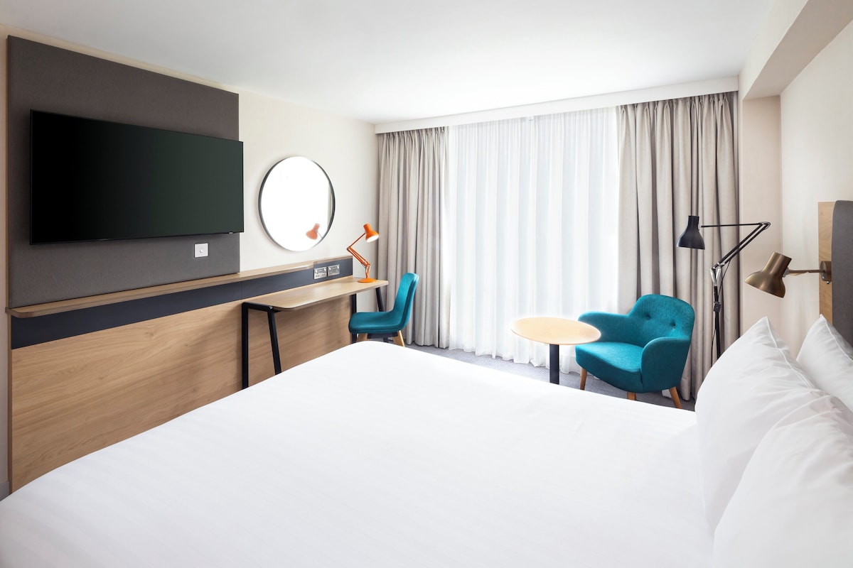 Habitación de hotel con cama, escritorio, silla, televisor y ventana con cortinas.