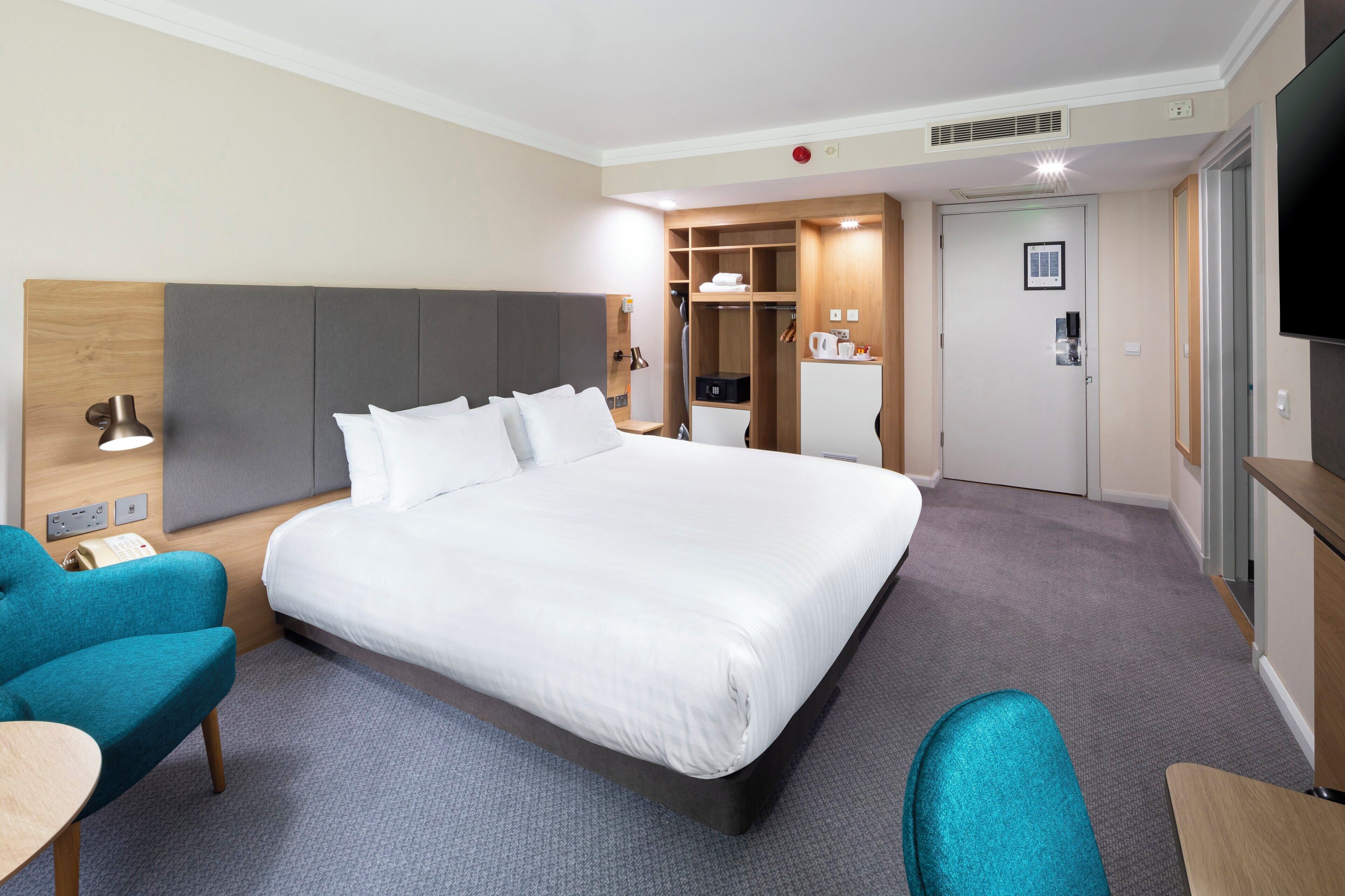 A hotel room with a large bed, a desk, a chair, a TV, and a wardrobe.