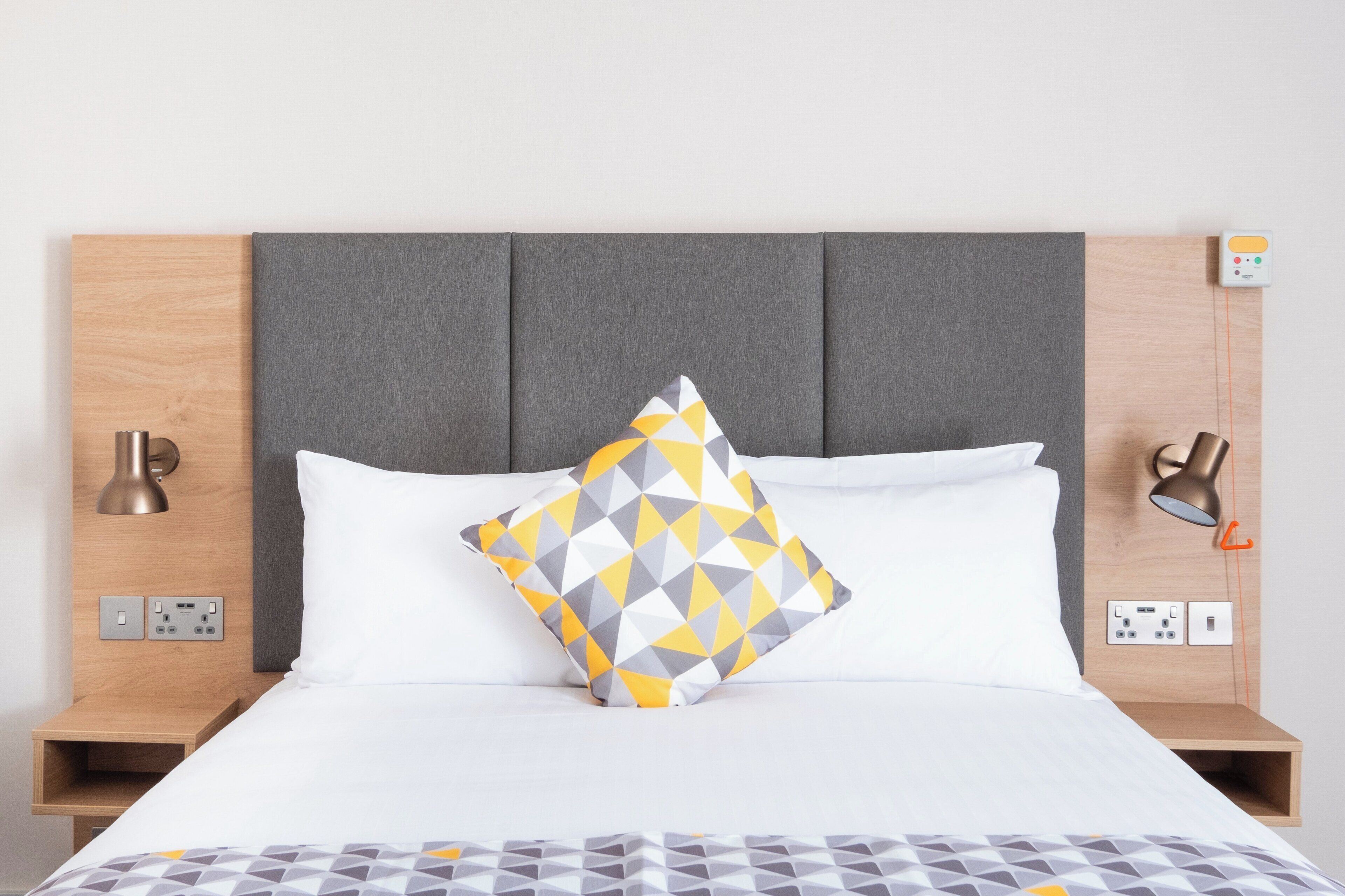 A neatly made bed with a geometric patterned pillow, a grey headboard, bedside lamps, and a power outlet on the nightstand.