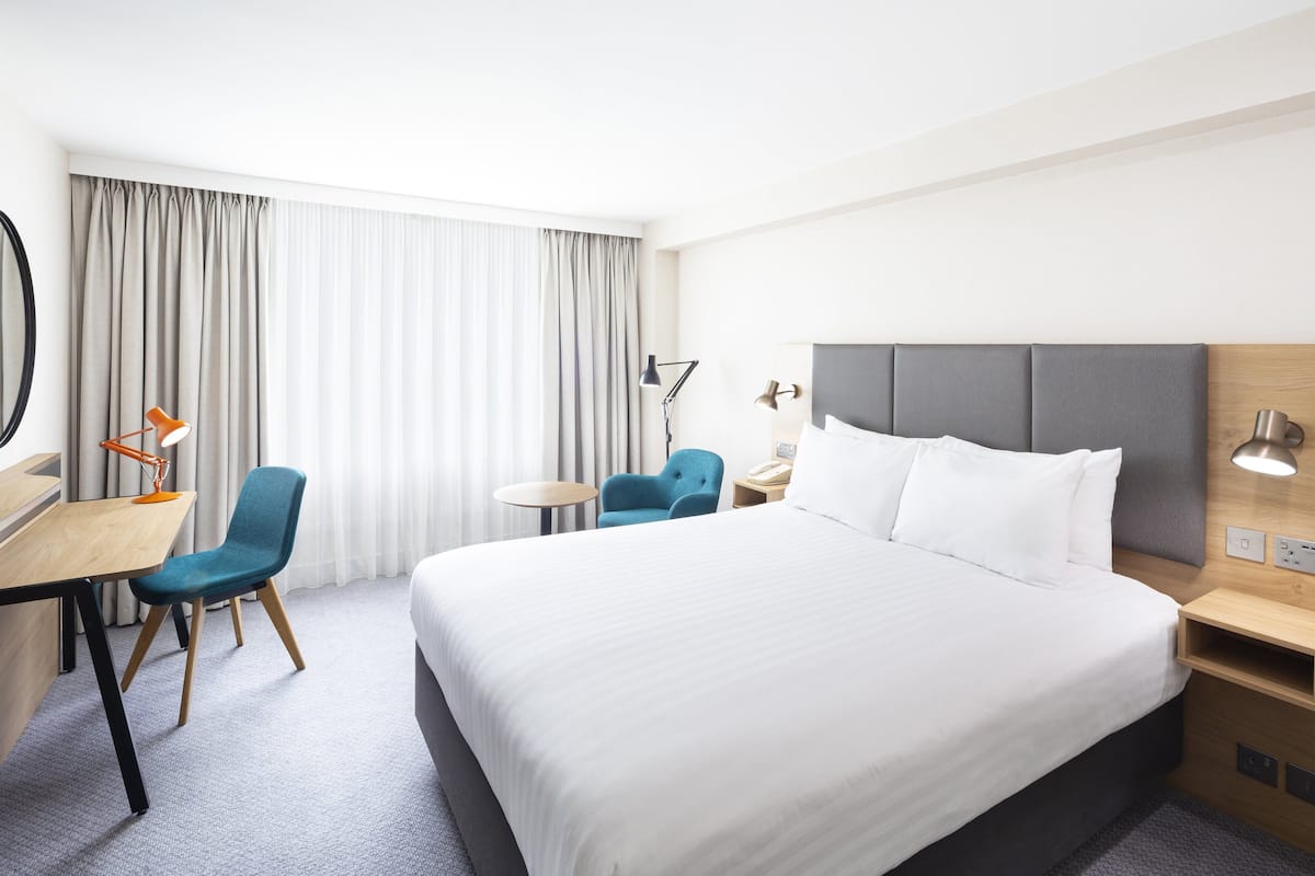 A hotel room with a large bed, a desk with a chair, a small table, and a lamp.