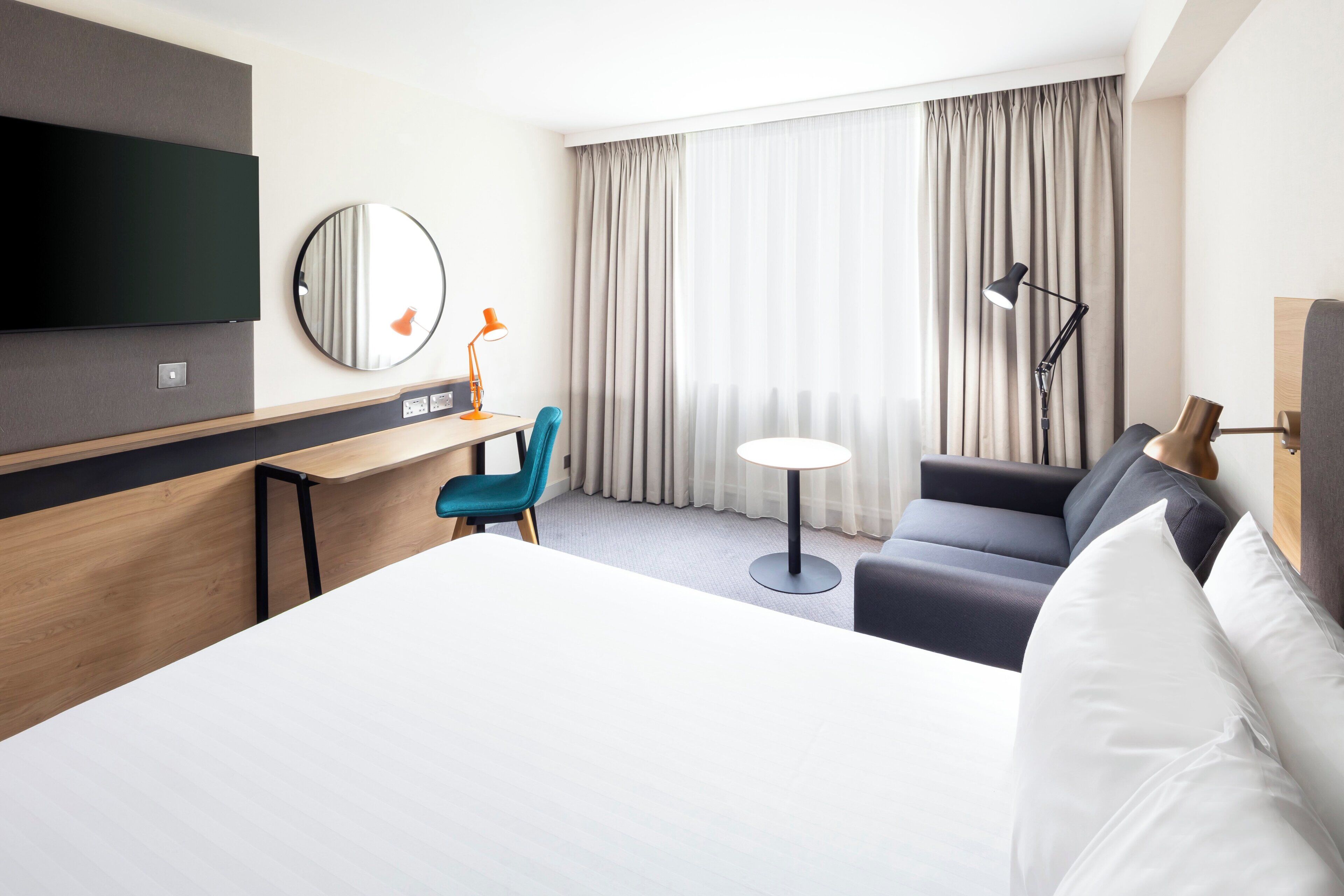 Una habitación de hotel con cama, escritorio, silla, televisor y ventana con cortinas.
