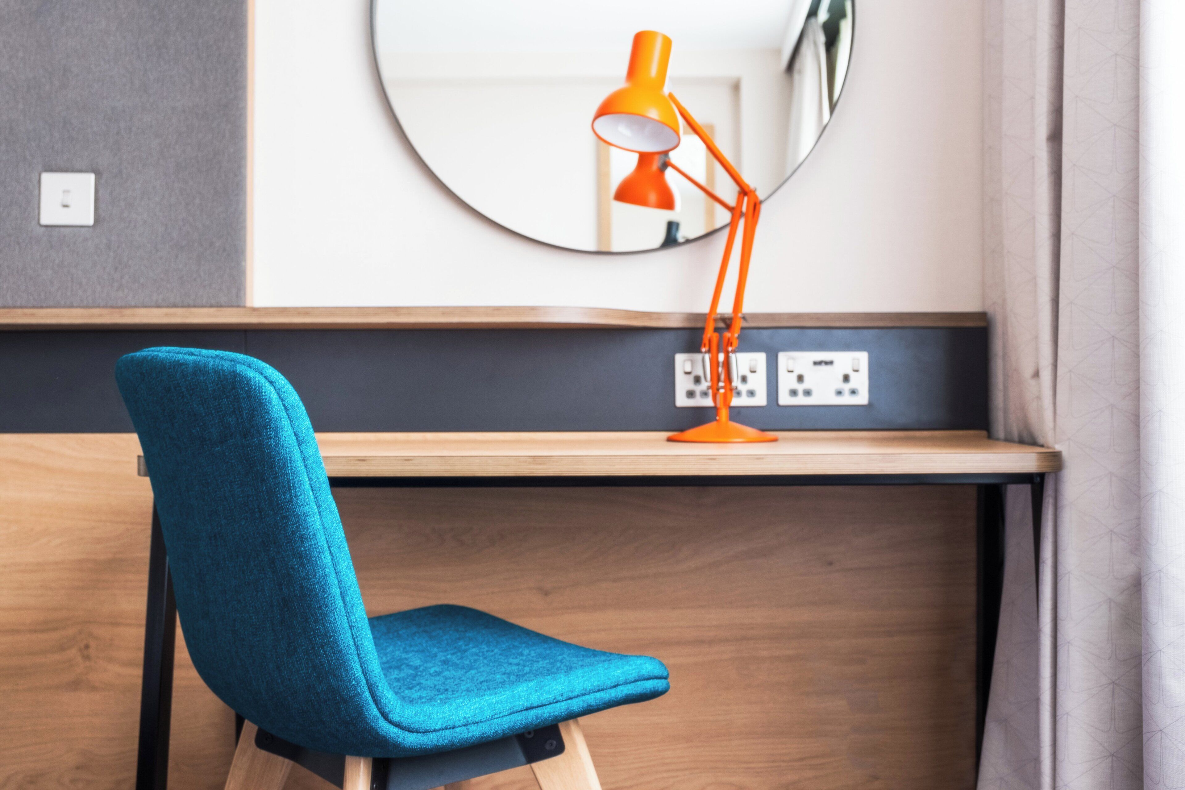Une chaise bleue et une lampe de bureau orange sont posées sur un bureau en bois, au-dessus duquel se trouve un miroir.