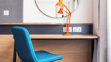Une chaise bleue et une lampe de bureau orange sont posées sur un bureau en bois, au-dessus duquel se trouve un miroir.