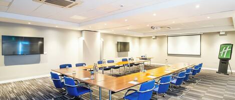 A conference room with a long table, blue chairs, a projector screen, and a flat-screen TV on the wall.