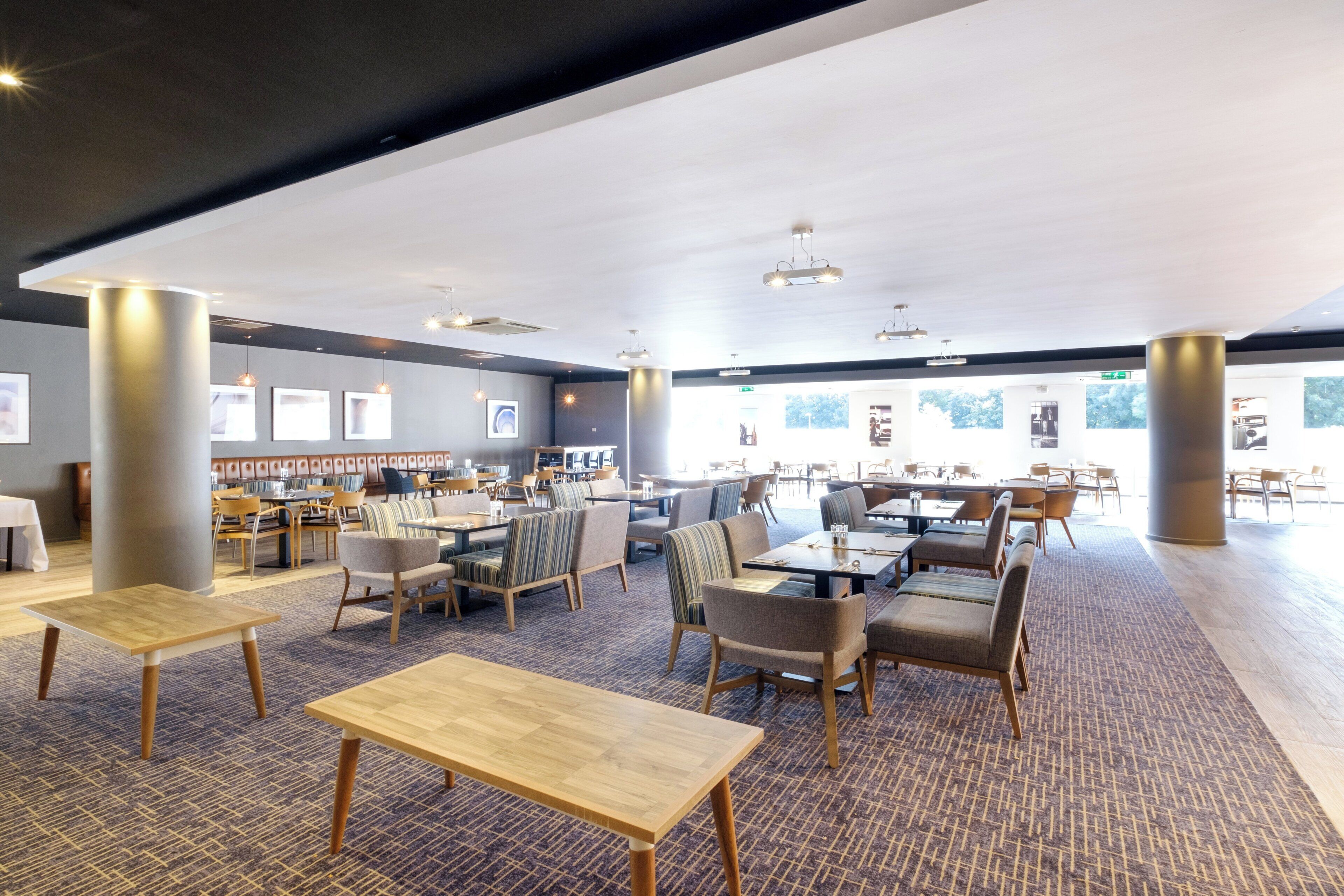 A spacious dining area with multiple tables and chairs, large windows, and a high ceiling with recessed lighting.