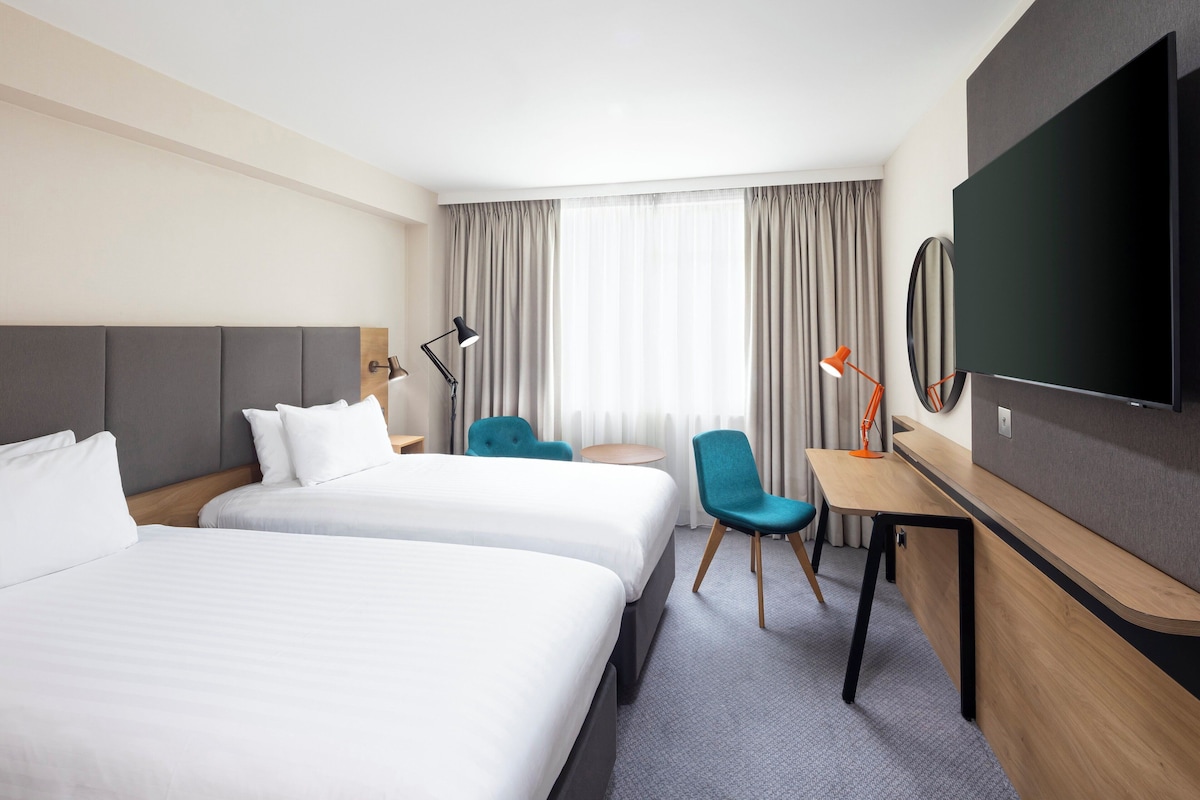 Habitación de hotel con dos camas, un escritorio, una silla, un televisor y una ventana con cortinas.