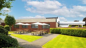 A courtyard with picnic tables and umbrellas, a brick building, and a hedge. - Holiday Inn Coventry M6, Jct.2 by IHG (Coventry)