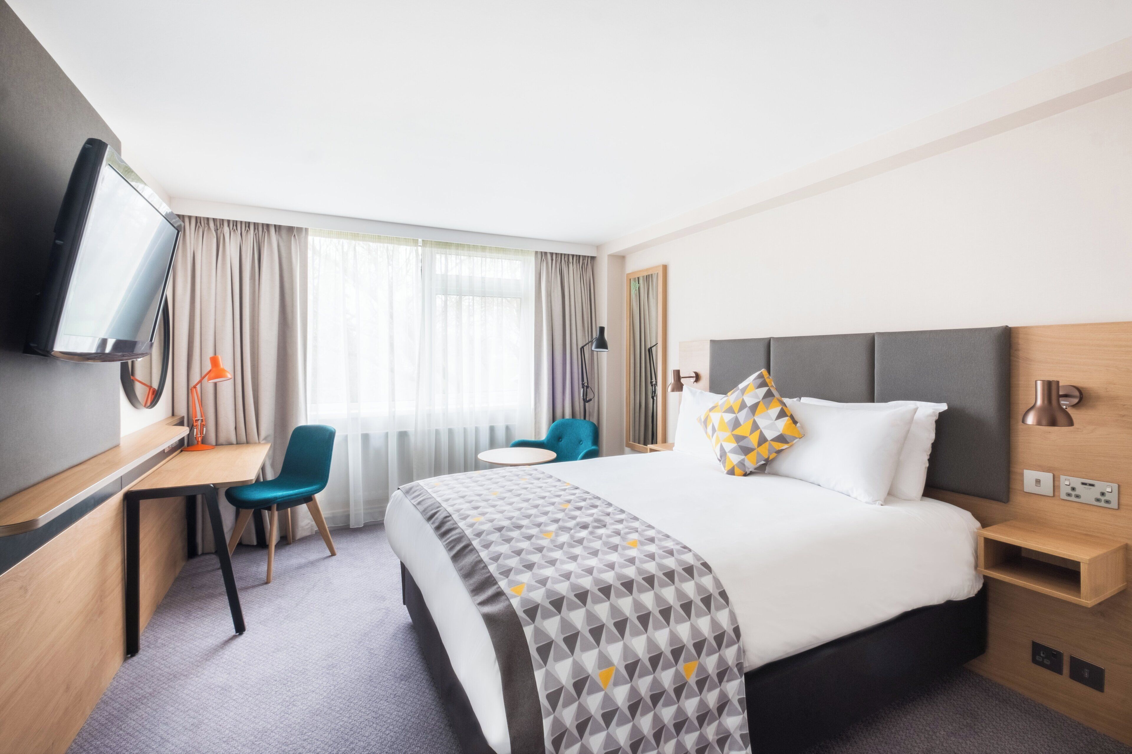 A hotel room with a bed, desk, chair, TV, and a window with curtains.