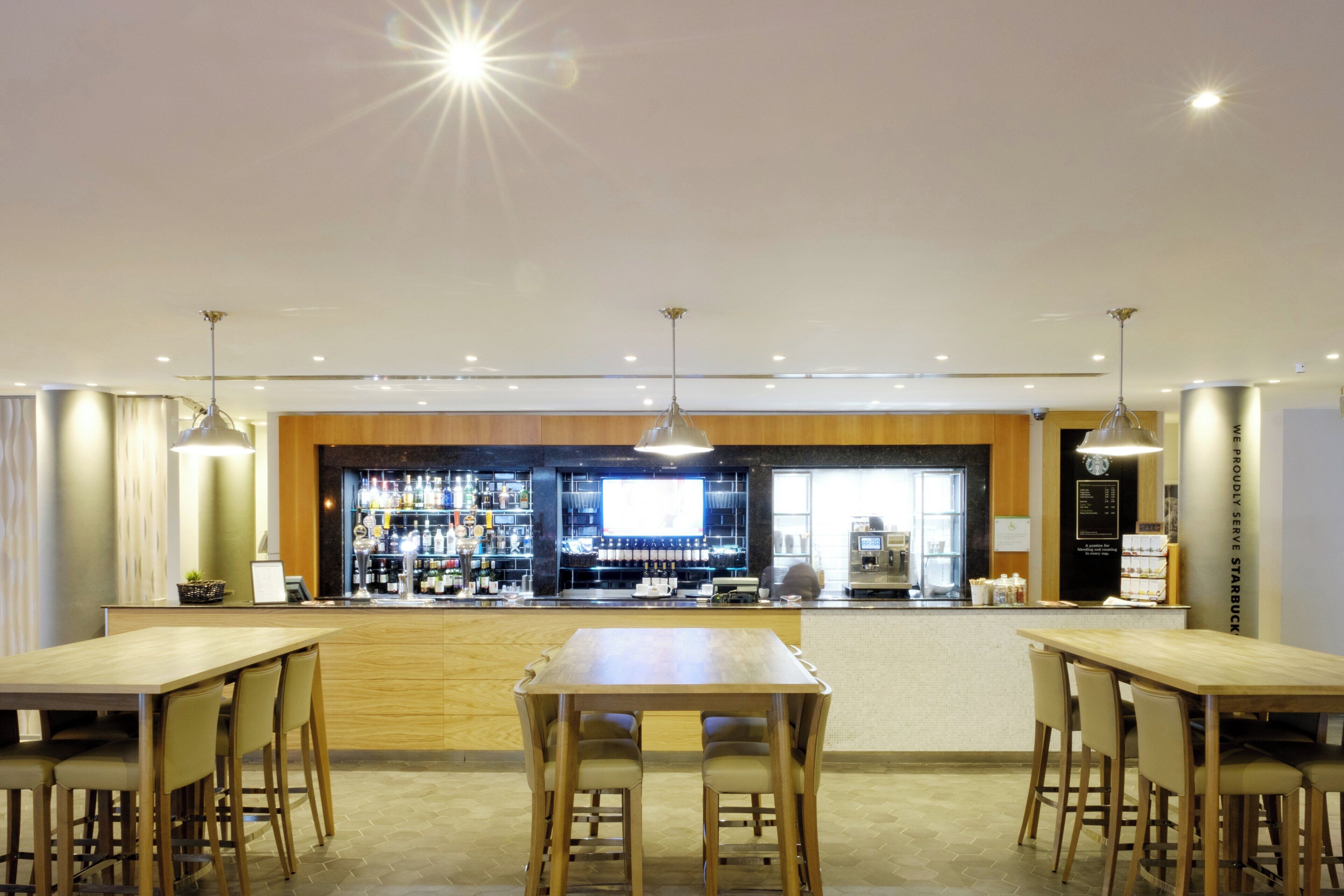 A modern bar area with wooden tables and chairs, a counter with a variety of bottles, and a large screen.