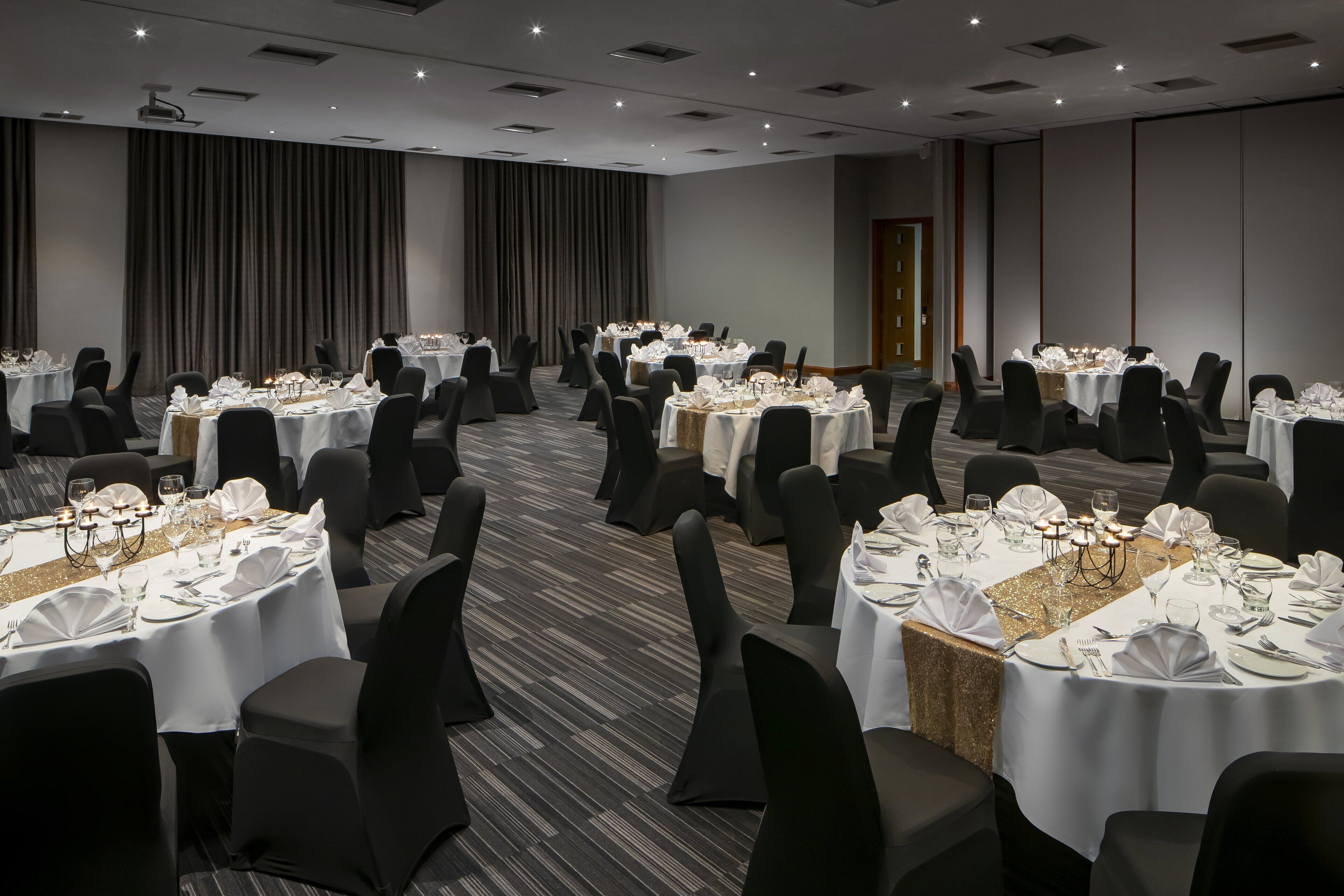 A banquet hall with multiple round tables set for dining, each with white tablecloths and black chairs.