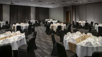 A banquet hall with multiple round tables set for dining, each with white tablecloths and black chairs.