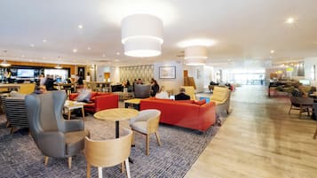 Un amplio vestíbulo de hotel con varias zonas de estar, incluyendo sofás y sillones, y una zona de bar al fondo.
