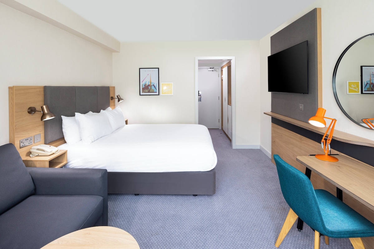 Habitación de hotel con cama, escritorio, silla, televisor y teléfono en la mesita de noche.
